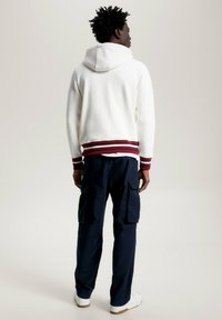 Weißer Hoodie mit rotem und marineblauem gestreiften Saum, kombiniert mit einer marineblauen Cargo-Hose. Das Outfit hat eine lockere Passform und minimale Details.