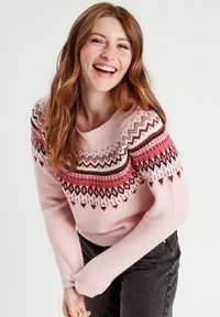 Różowy sweter z dzianiny z kolorowym wzorem fair isle w czerwonym, białym i czarnym kolorze. Miękka tekstura i okrągły dekolt. Na sobie ciemne dżinsy.