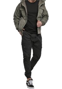 Kurtka typu bomber w kolorze oliwkowej zieleni z wieloma kieszeniami, czarny T-shirt, czarne spodnie typu cargo ze ściągaczami oraz czarne sneakersy z białymi podeszwami.