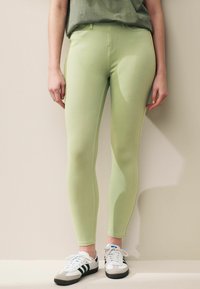 Hellgrüne, sportlich geschnittene Leggings mit glatter Textur und hohem Bund. Kombiniert mit weißen Sneakers mit schwarzen Akzenten und einem grauen Oberteil.