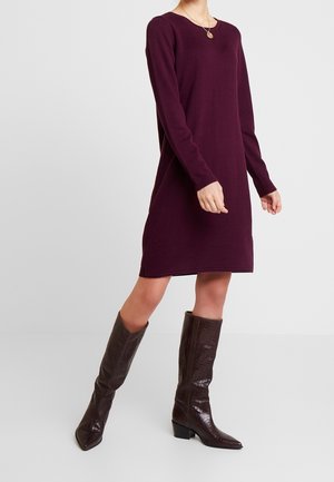 Femme portant une robe prune mi-longue à manches longues avec des bottes hautes texturées marron foncé sur un fond clair uni.