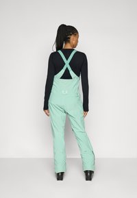 Monos de nieve de color verde menta con tirantes cruzados, cierres ajustables y bolsillos con cremallera, combinados con una camiseta de manga larga negra ceñida.