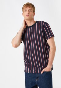 Camiseta a rayas en azul marino con rayas verticales rojas y blancas, cuello redondo, mangas cortas y corte relajado. Combinada con pantalones de mezclilla.