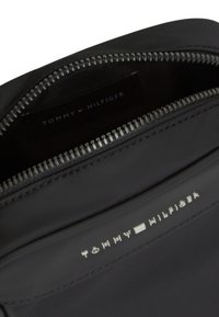 Μαύρη τσάντα Tommy Hilfiger με ασημένιο φερμουάρ μισάνοιχτο, που δείχνει το λογότυπο της μάρκας στο εσωτερικό και στην εξωτερική μπροστινή τσέπη.