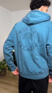 Sweat à capuche bleu avec un logo circulaire représentant un arbre et le texte "BLK VIS EST. 2016." Le tissu semble doux et légèrement texturé.
