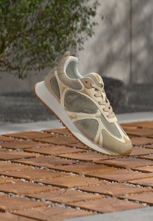 Valentino tenisica u bež i smeđim tonovima, s detaljima od mrežaste tkanine i ante, bijeli potplat, prikazana u zraku iznad terakota pločica na otvorenom.