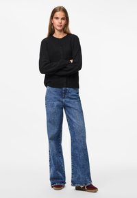 Cardigan noir en tricot avec un col rond et une fermeture à boutons, porté avec un jean large bleu et des baskets bordeaux.