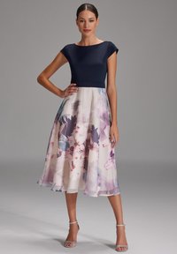Une robe bustier marine avec une jupe plissée à fleurs dans des couleurs pastel, présentant une silhouette en A et des sandales argentées à bride chevillère.