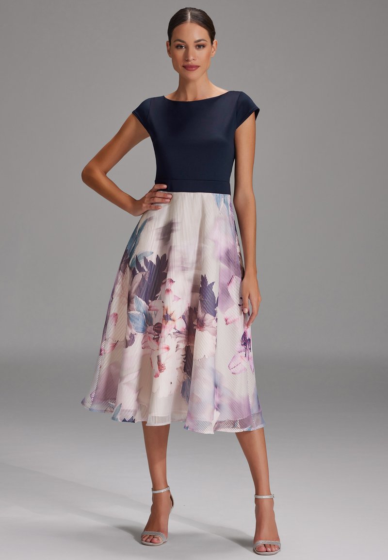 Une robe bustier marine avec une jupe plissée à fleurs dans des couleurs pastel, présentant une silhouette en A et des sandales argentées à bride chevillère.