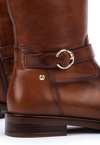 Bruine leren laars tot de knie met een bandje met een gouden ronde gesp, een zijrits en een lage gestapelde hak. Gladde textuur met stiksels.