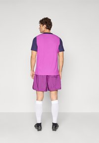 Lilla idretts skjorte med marineblå ermer, matchende lilla shorts, hvite sokker, og svarte fotballsko. Teksturert stoff med en avslappet passform.