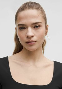 Kratak fokus na osobu s dugom kosom, obučenu u crni odjevni predmet s kvadratnim izrezom. Koža ima glatku teksturu, a oči su bistre i usredotočene.