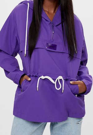 Femme portant un coupe-vent violet à capuche avec des cordons blancs et les mains dans les poches avant, associée à un jean bleu clair déchiré.