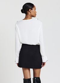 Blusa bianca con maniche a campana, caratterizzata da dettagli neri ai polsini; abbinata a una gonna nera aderente. Si vede la schiena della modella.