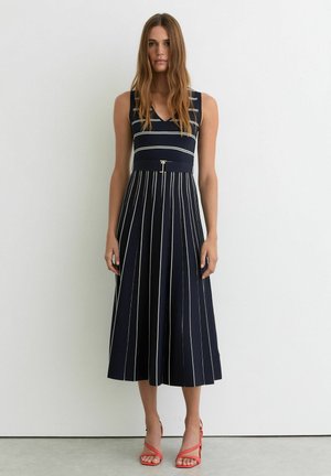 Nainen, jolla on yllään hihaton laivastonsininen mekko, jossa on valkoisia pystysuoria ja vaakasuoria raitoja, vyötärökoristelu ja punaiset korkeakorkoiset sandaalit, joissa on remmit.