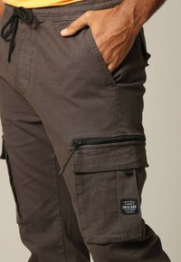 Cargobroek van donkerbruin stof, met een elastische tailleband, zijzakken en gevoerde cargozakken met rits. Inclusief logo-opdruk.