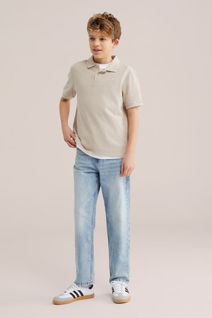 Fiú áll, bézs színű galléros pólót, világoskék farmert és fehér, fekete csíkos sportcipőt visel egyszínű háttér előtt.