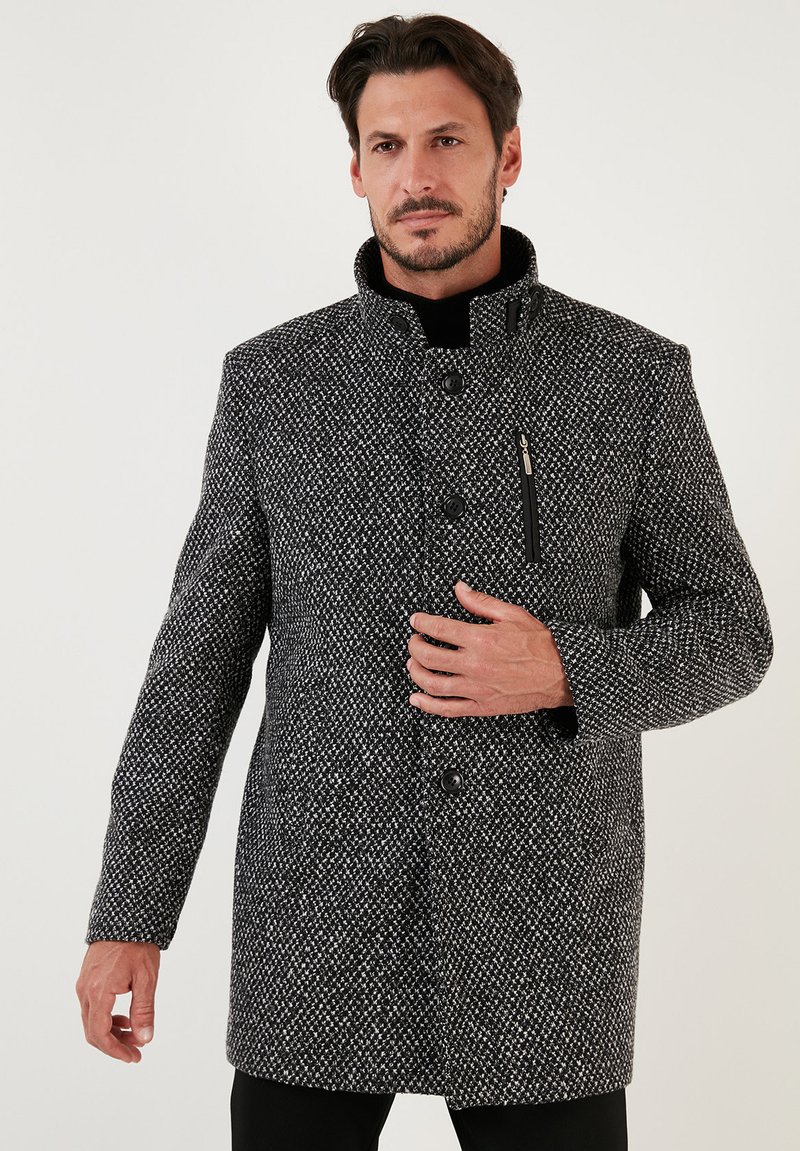 Abrigo de hombre en patrón de tweed blanco y negro; cuenta con un cuello alzado, botones frontales y un bolsillo lateral con cremallera. Textura suave, ajuste entallado.