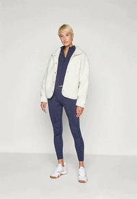 Osoba ubrana w granatowy zestaw sportowy z zamkiem błyskawicznym, legginsy, białą puszystą kurtkę i białe sneakersy, stojąca na tle jednolitej białej ściany.