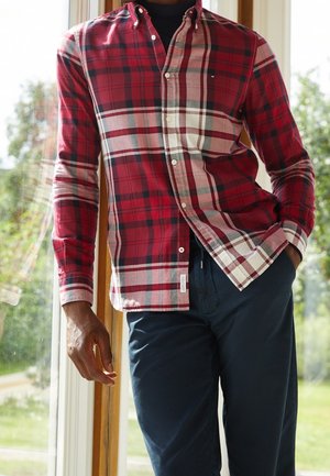 Mann trägt ein rot-weiß kariertes Hemd über einem schwarzen Unterhemd und marineblaue Hose, steht nahe einem Fenster mit Grünpflanzen draußen.