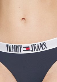 Marinblå bikiniunderdel med ett vitt elastiskt midjeband som har "TOMMY JEANS" i svart och röd text, slät yta, låg midja.