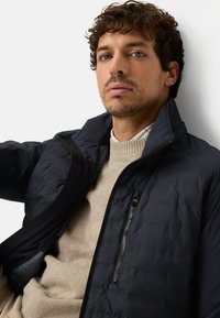 Marineblauwe bomberjas met een zak met rits en een hoge kraag, gedragen over een beige gebreide trui. Gladde stof met een matte afwerking.