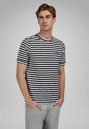 Junger Mann mit hellbraunen Haaren, der ein schwarz-weiß gestreiftes T-Shirt und hellgraue Jeans trägt, die Hände in den Taschen, blickt nach rechts.