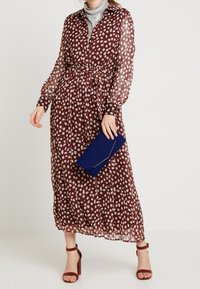 Vestido largo marrón con lunares blancos, con cierre de botones en la parte delantera, cintura ajustada, tejido transparente, combinado con un bolso azul de mano y sandalias marrones de tacón cuadrado.