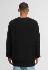 Maglione nero lavorato a maglia con un motivo testurizzato, maniche allungate e polsini a costine. Sul retro presenta sottili accentuazioni sulle spalle.