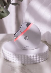 Weißes, rundes Reinigungsgerät mit reflektierendem silbernen Oberteil und rosa Akzenten. Es steht auf einer strukturierten weißen Basis mit kreisförmigen Mustern.
