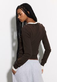 Braunes langärmliges Oberteil mit figurbetontem Design, cremefarbenen Akzenten an den Bündchen und strukturiertem Stoff. Modell trägt graue Jogginghose, Ansicht von hinten.