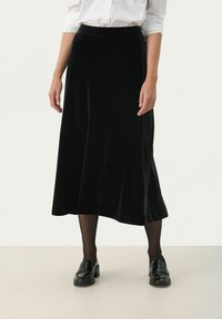 Schwarzer Samt-Midi-Rock mit glatter Textur, ausgestellter Form und nahtlosem Taillenbund, kombiniert mit schwarzen Slippern und Strumpfhosen.
