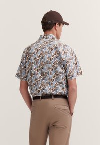 Kurzärmliges Hemd mit einem blau-braun-weißen Blumenmuster; aus Baumwolle; Rückansicht; kombiniert mit khakifarbenen Hosen.