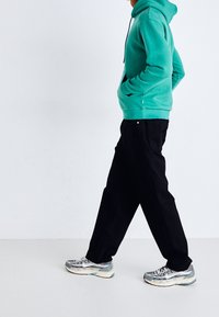 Turkos hoodie med framficka, svarta byxor med avslappnad passform och grå/vita träningsskor med blå detalj.