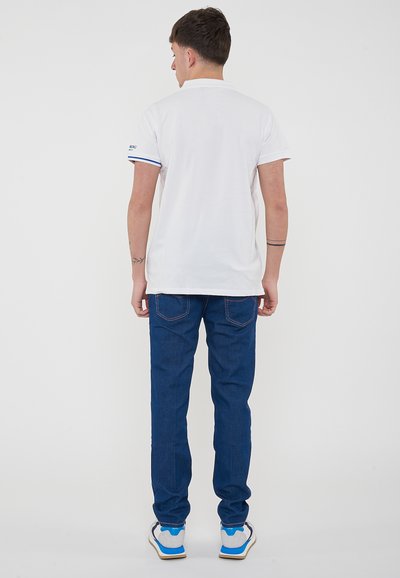 Polo shirt bianco a maniche corte con dettagli blu e colletto semplice, abbinato a jeans in denim blu con cuciture a contrasto e tasche posteriori.