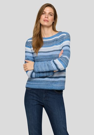 Frau mit langen hellbraunen Haaren, die einen blau-weiß gestreiften Strickpullover und dunkelblaue Jeans trägt, Arme verschränkt, neutraler Hintergrund.