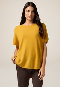 Suéter grande de punto en color mostaza con mangas cortas, cuello redondo y dobladillo acanalado, combinado con pantalones marrón oscuro.