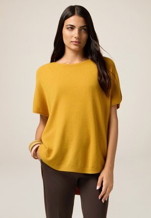 Gemusterter, senfgelber, übergroßer Pullover mit kurzen Ärmeln, rundem Ausschnitt und geripptem Saum, kombiniert mit dunkelbraunen Hosen.