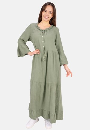 Frau lächelt, trägt ein langes, hellgrünes, geknöpftes Kleid mit Rüschenkragen und weiten Ärmeln, kombiniert mit weißen Sneakers, stehend.