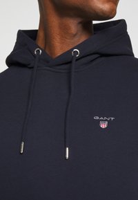 Primo piano di una persona che indossa un hoodie GANT blu navy con stringhe e un piccolo logo ricamato sul petto.