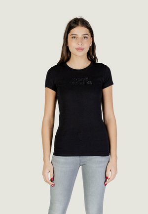 Joven con cabello largo y castaño, vistiendo una camiseta negra ajustada y jeans gris claro, posando frente a un fondo claro y sencillo.