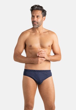Homme torse nu aux cheveux courts et foncés avec une barbe, portant un slip bleu marine à petits motifs rouges, debout devant un fond blanc.