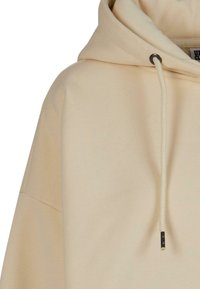 Felpa beige in cotone con cappuccio regolabile, vestibilità over size, texture a coste e occhielli in metallo argentato. Presenta dettagli di design minimalista.