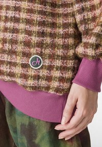 Pulover tricotat dintr-un material bouclé multicolor, cu o textură modelată, tiv violet cu nervuri și un mic logo circular pe față.