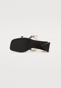 Sandalia negra y metálica con un tacón triangular. Presenta una tira suave, suela texturizada y un diseño minimalista.