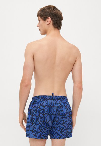 Ung man med kort brunt hår står vänd bort, iklädd blå och svarta shorts med ett upprepande mönster av "DSQUARED2"-logotypen.