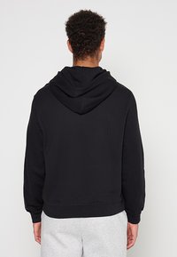 Czarna bluza z kapturem o teksturowanej powierzchni, wyposażona w kaptur ze sznurkiem i ściągacz na dole. Tył ukazuje rękawy sięgające nadgarstków.