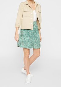 Světle béžová zkrácená bunda s velkými knoflíky a dvěma náprsními kapsami; sukně s zeleným květinovým potiskem; bílé tenisky.