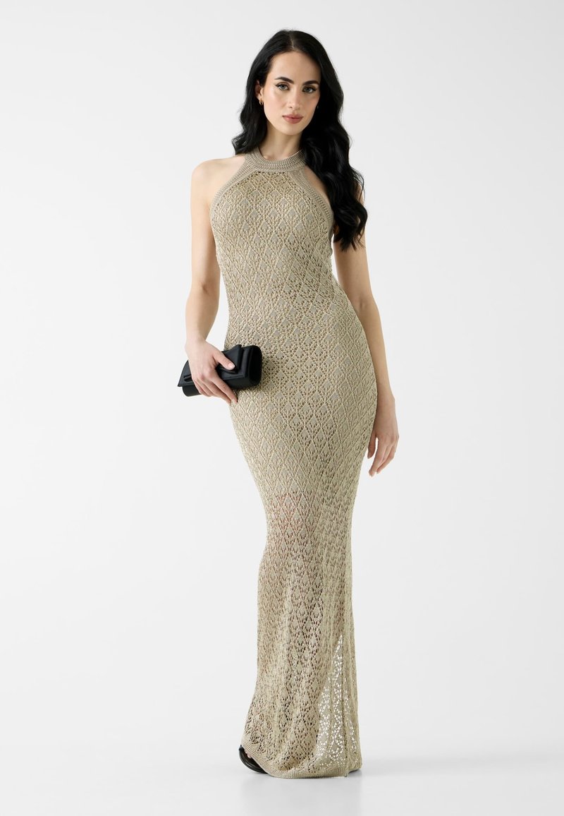 Femme aux longs cheveux foncés portant une robe longue beige en crochet avec un col halter, tenant une petite pochette noire sur un fond blanc.