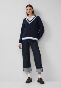 Marineblauwe v-hals trui met een lichtblauwe en witte gestreepte kraag en zoom, gecombineerd met donkere denim wijde broek en bruine loafers met witte sokken.